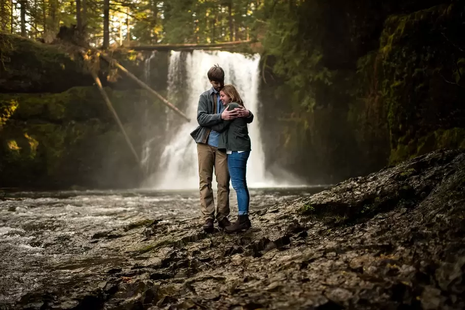 ​Winter Waterfall Engagement Photos from Photographer Robert Knapp, a couple holds each other standing in the mist given off from a giant waterfall. All is dark around. Light seems to be coming from the water itself 