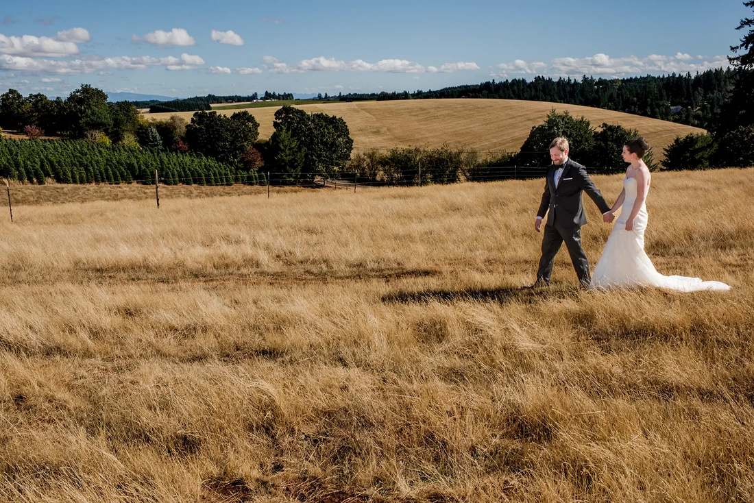 Farm Wedding Oregon Rustic ​Chic Style with Robert Knapp Photographer A bride and groom in tuxedo and wedding dress with a long train and no straps, walk across a brown dry field of grasses. In the distance the horizon of rolling hills meets a light blue sky and a thin line of white puffy clouds. 