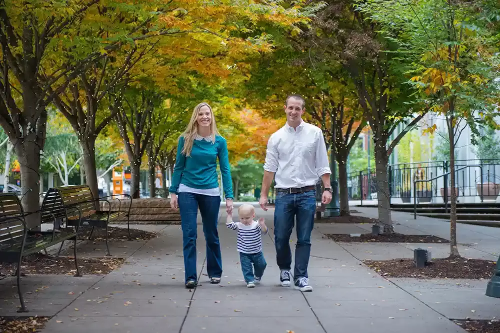 Family photography by robert knapp of a family walking on a sidewalk. holiday lights wrap the trees and brances, they glow dimly. The trees are starting to tun color for autum. 