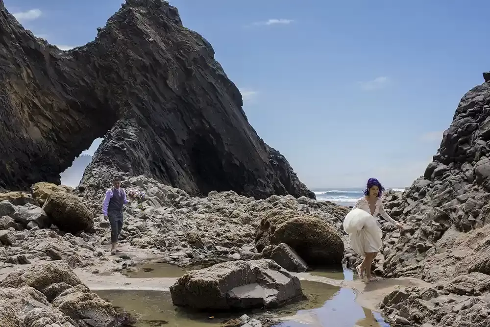 Photographers Portland at bridal shoot, a man and a woman in formal wedding attire, of suit and white wedding dress walk across a stone covered shore, the ocean in the background as a giant stone arch moves from the cliff to the ocean