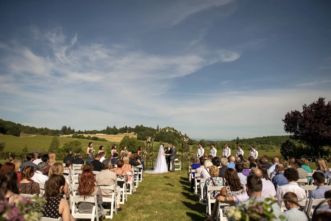 ​Vineyard Wedding Oregon Vineyard Wedding Oregon with rolling hills in the distance and wispy clouds overhead. Guests lean int to see the ceremony. 