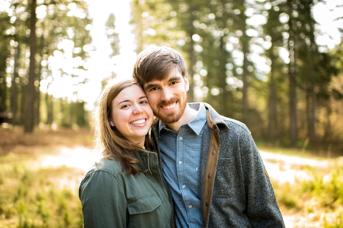 ​Winter Waterfall Engagement Photos from Photographer Robert Knapp a couple looks to the camera smiling cheek to cheek. The sun shines at them from behind 
