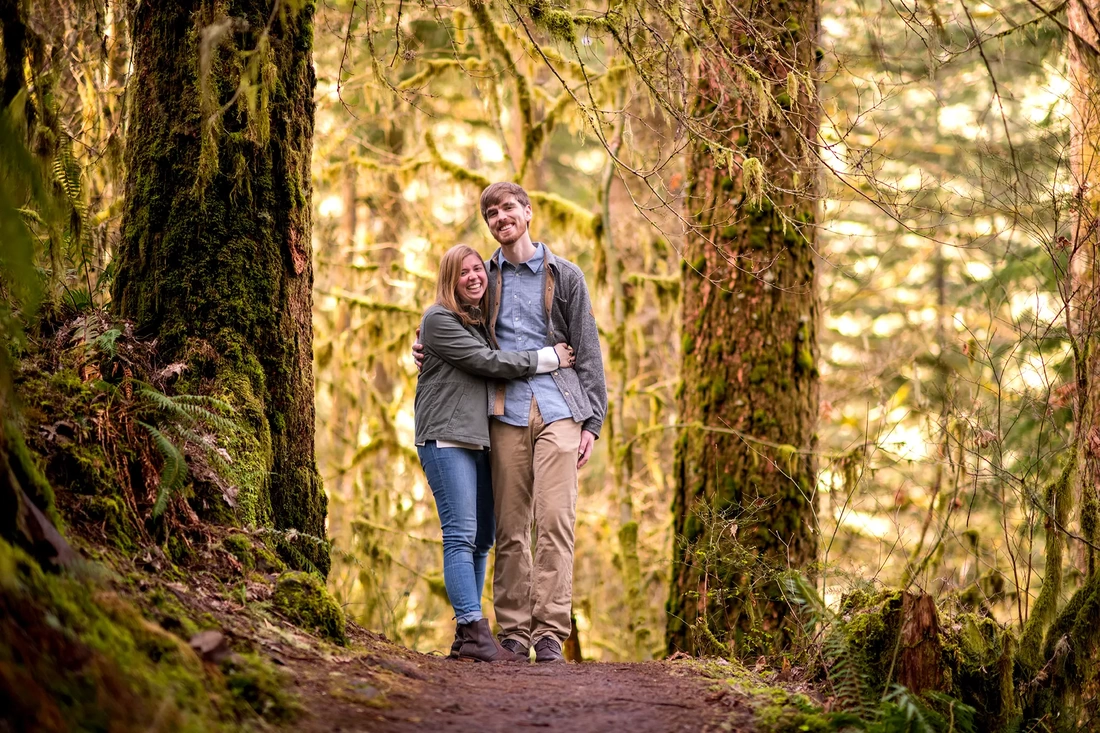 ​Winter Waterfall Engagement Photos from Photographer Robert Knapp a couple stands on a path and smiles as they look to the camera. The forest is lit with golden light around them 