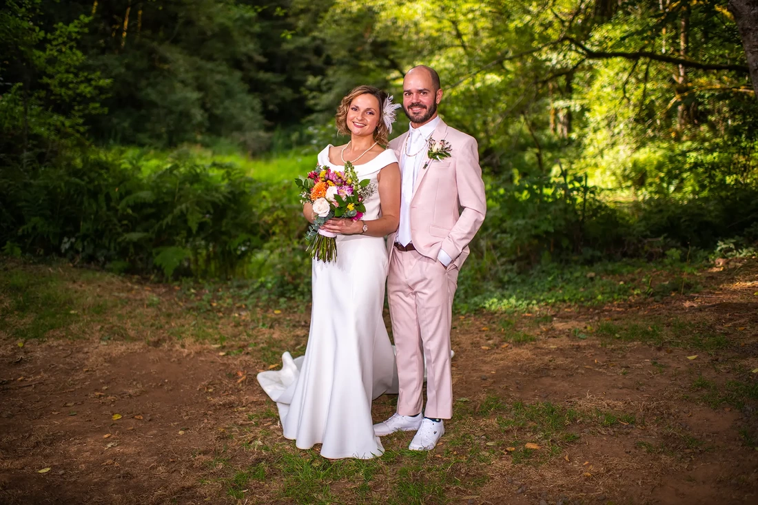 A bride and groom stand in a forest setting, white dress and pink suit. A Beautifully lit Horning's Hideout Wedding; Modern Art Photograph