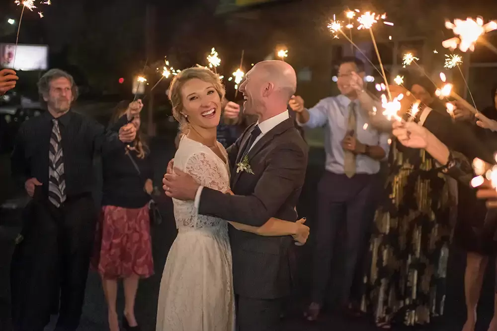 Portland Wedding Photographer Robert Knapp captures a photo of a couple holding each other after their wedding reception as their guests light sparklers around them in a circle. 