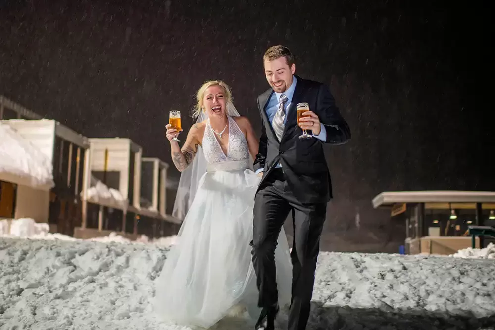Portland Wedding Photographer Robert Knapp Photographs this bride and groom running in the snow with their beers. They are very happy. It is snowing. It is after dark. No flash was used. The photo is well lit. 