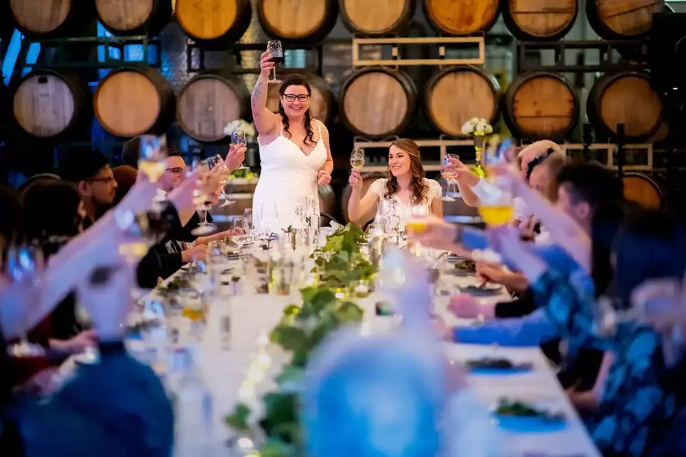Portland Wedding Photographer Robert Knapp Pointing a camera across a very long reception table where all the guests are seated. At the head of the table two raise a glass. All the guests follow suit. Wine Barrels make a great background to the scene 