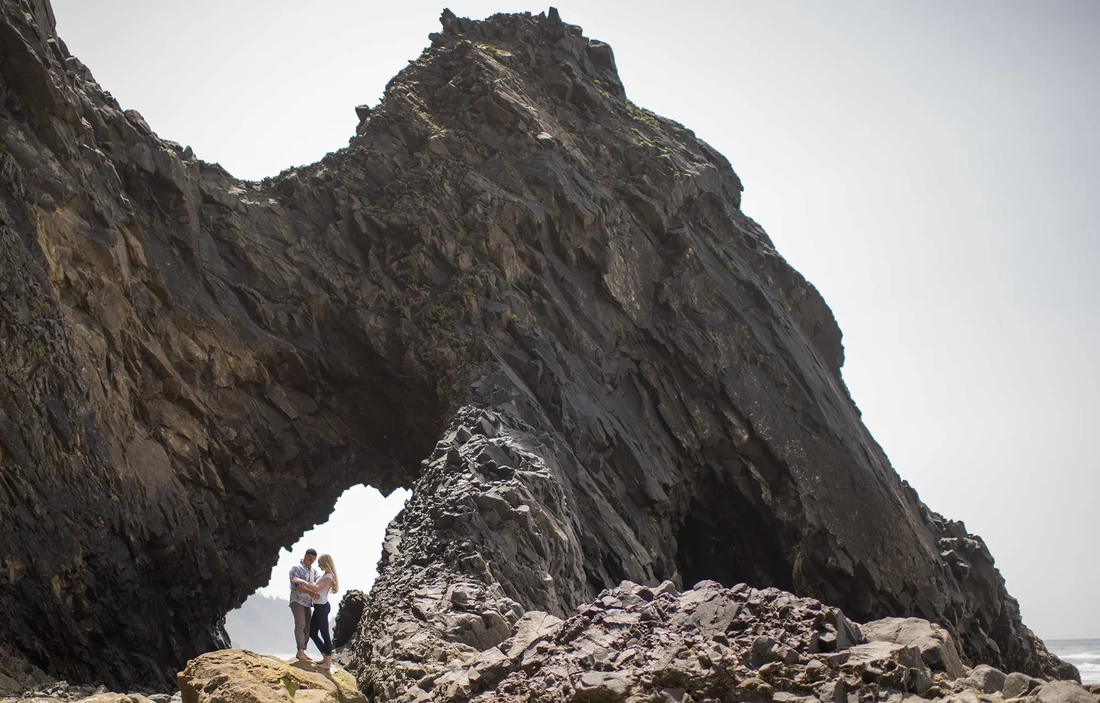 Cannon Beach Engagement Photos, a couple stands on the beach under an interesting rock ledge, the ocean is only just in the frame