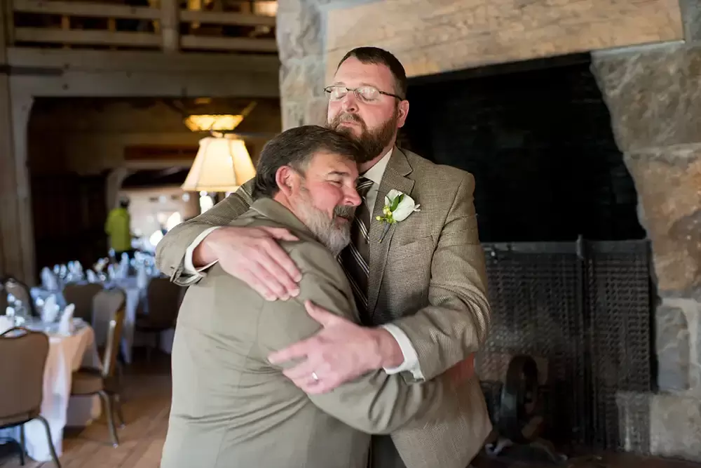 Wedding at Timberline Lodge groom hugging father both with eyes closed in front of a Gian fireplace