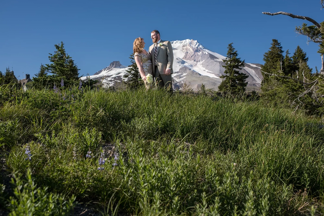 Wedding at Timberline Lodge ​from photographer Robert Knapp Bride and groom stand in a field of wildflowers with mount hood in the distance. The bride and groom look to each other before a Wedding at Timberline Lodge