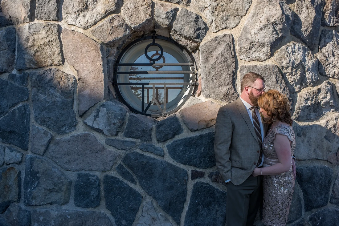 Wedding at Timberline Lodge, sunset begins and the shadows grow long as a groom kisses his bride on the top of the head. The masonry of the lodge is the background