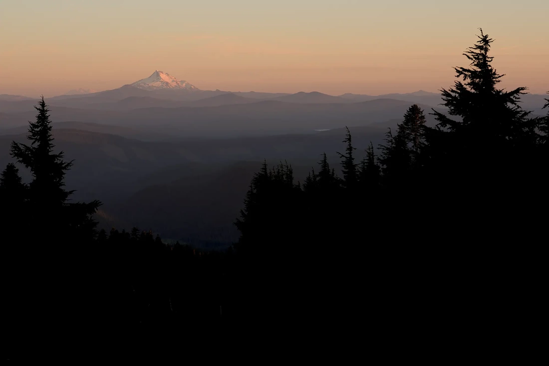 Wedding at Timberline Lodge comes to an end. Sunset over the Cascade Range Mount Jefferson on the horizon in a low orange light of sunset. Treetops in the foreground are black outlines of conifer trees. 