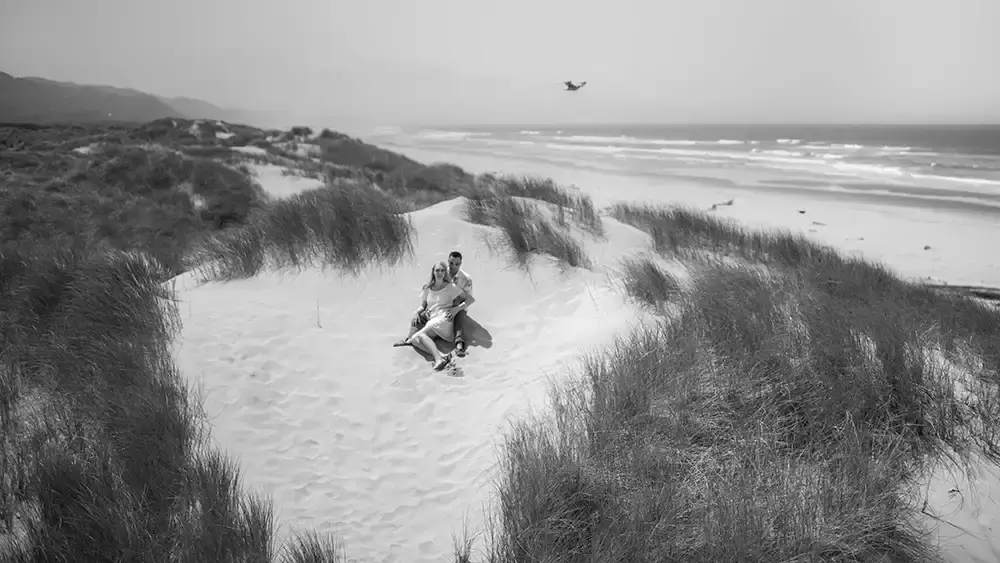 A couple sits in a sand dune near the ocean and a seagull is just overhead Cannon Beach Engagement Photos With Photographer Robert Knapp