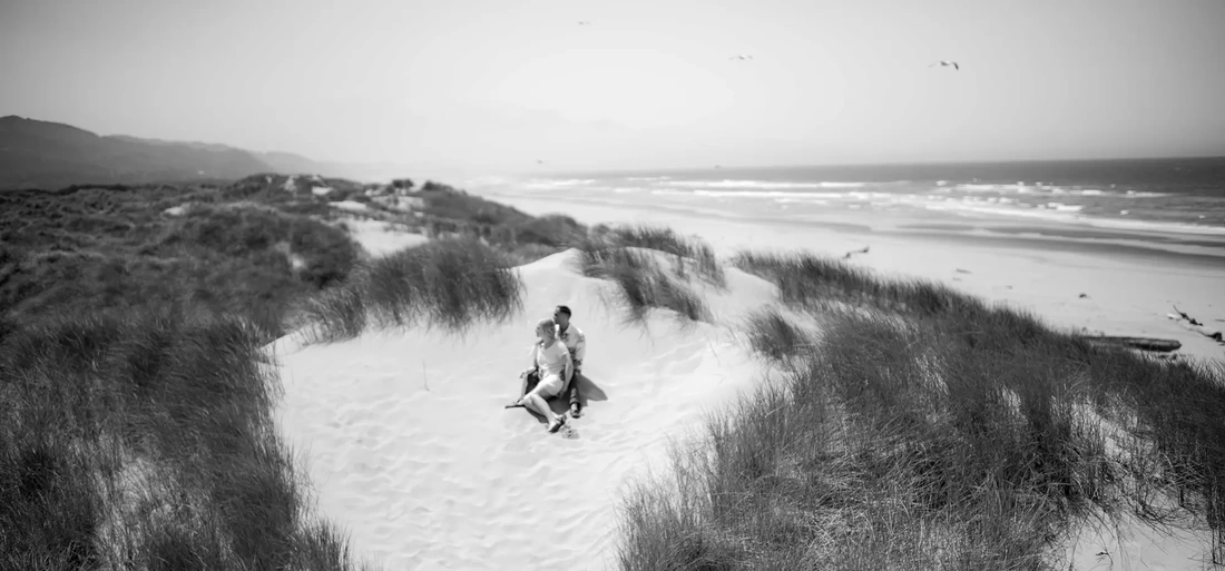 Cannon Beach Engagement Photos a couple sit on a sand dune, the ocean is in the background, seagulls fly overhead