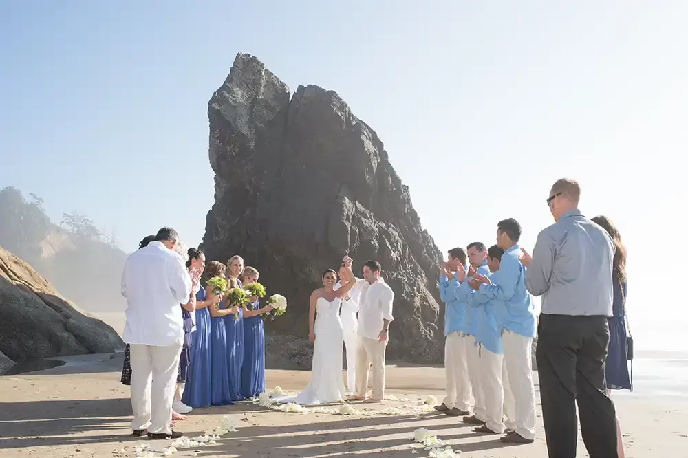 Wedding on the Beach
Cannon Beach Wedding
Photographer Robert Knapp all clap as the ceremony completes, bride and groom hold hands together over their head. A jagged cliff rises out of the sand behind them