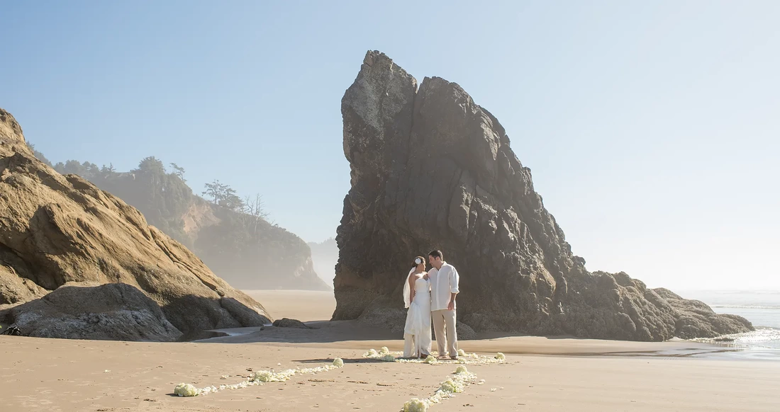 Wedding on the Beach Cannon Beach Wedding Photographer Robert Knapp Bride and Groom stand in front of a massive rock between the ocean and the cliffs. This is their ceremony site on the beach