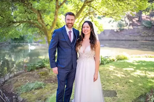 Portland Wedding Photographer Robert Knapp bride and groom summer wedding standing int the shade. A pond behind them, a goose swims in the pond. The groom is in a blue suit, the bride is in an off white wedding dress, they look to the camera with great smiles. 