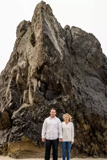 Oregon Coast Engagement Photos a couple hold hands in front of a larger stone sticking out of the beach. The wind blows the woman's hair perfectly