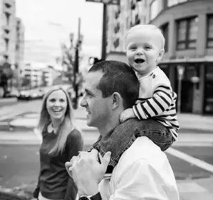 A family of three, mother father and baby stand at an intersection waiting to cross the road. Baby looks to the camera with a excited smile. Father looks to the traffic, Mother looks to the baby with great happiness