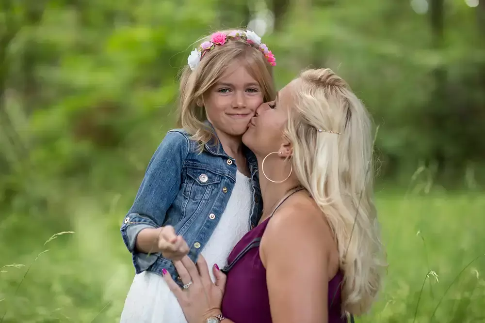 ​daughter looks to the camera, mother kisses the cheek of the daughter Garden Photoshoot with Robert Knapp one of the highly sought after Family Photographers Portland has to offer