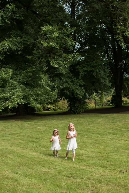 two little girls walk across a great lawn bordered by trees Garden Photoshoot with Robert Knapp one of the highly sought after Family Photographers Portland has to offer