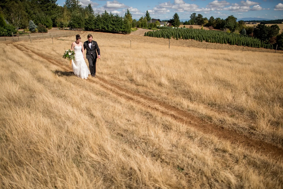 Farm Wedding Oregon Rustic ​Chic Style with Robert Knapp Photographer Mid day, bright blue sky, little puffy white clouds dot the horizon, bride and groom walk a path in a field. They wear their formal wedding dress and tux. 