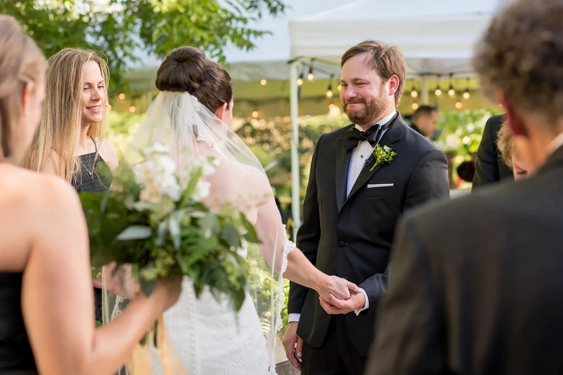 Farm Wedding Oregon Rustic ​Chic Style with Robert Knapp Photographer The ceremony photographed through crowd of people standing. The groom looks to his bride with a very full heart. In the background the reception tent is twinkling away. 