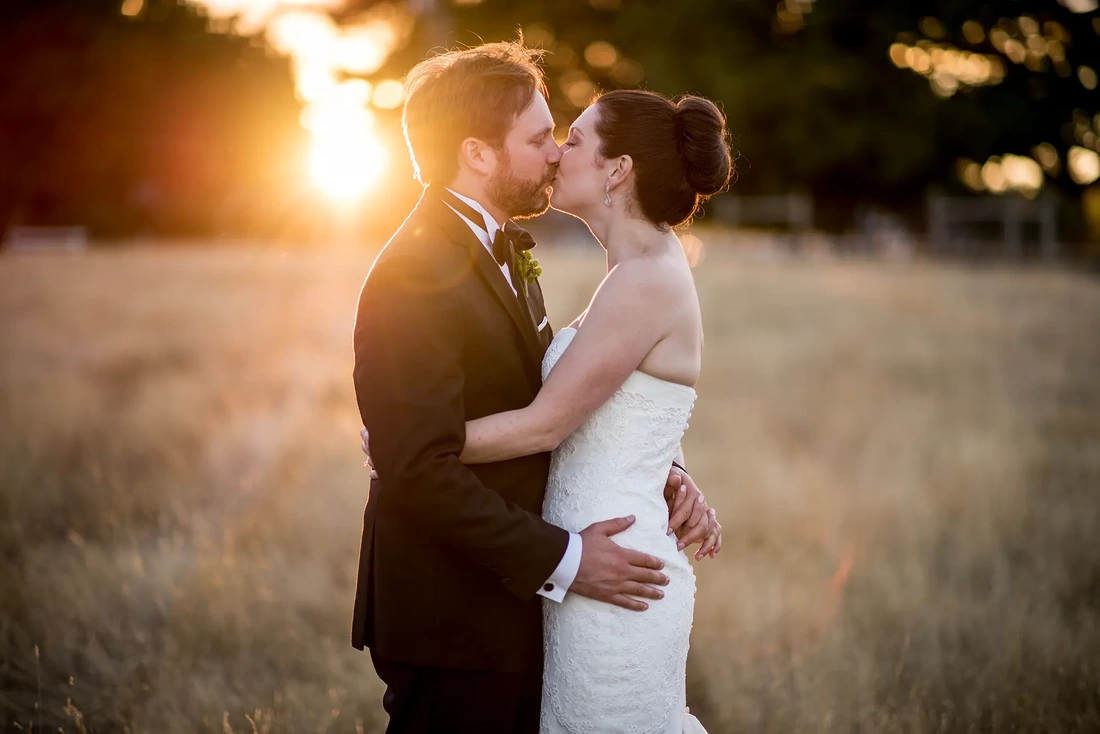 Farm Wedding Oregon Rustic ​Chic Style with Robert Knapp Photographer bride and groom kiss at sunset. amber grasses and golden sunset fill the frame. A solar flare circlets the shoulder of the groom fingers are woven together holding hands. 