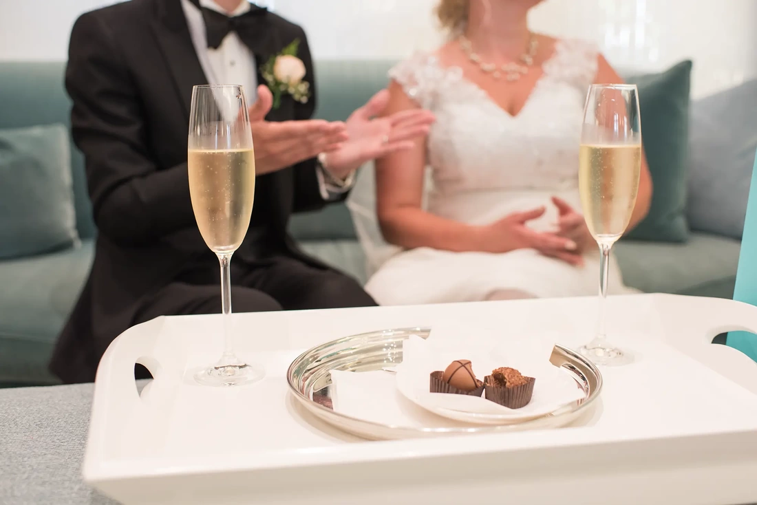 Hotel Deluxe Wedding in Portland Oregon by Photographer Robert Knapp in focus, two chocolates sit on a silver platter, gently out of focus two champagne glasses sit on a serving tray, dramatically out of focus bride and groom sit on a couch and talk with their hands