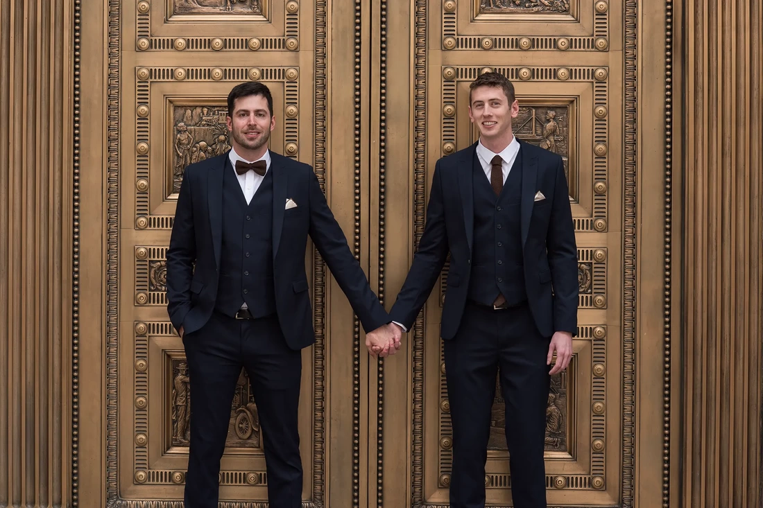 two groom hold hands in front of golden doors LGBT Wedding Photographer Robert Knapp at ​Tanner Springs Park in Portland Oregon