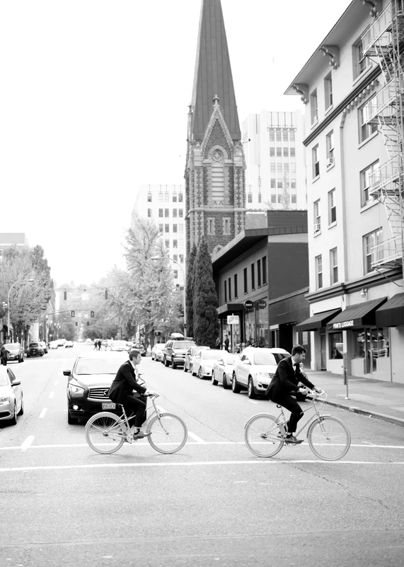 two guys gross the intersection on bicycles in front of a cathedral in black and white LGBT Wedding Photographer Robert Knapp at ​Tanner Springs Park in Portland Oregon