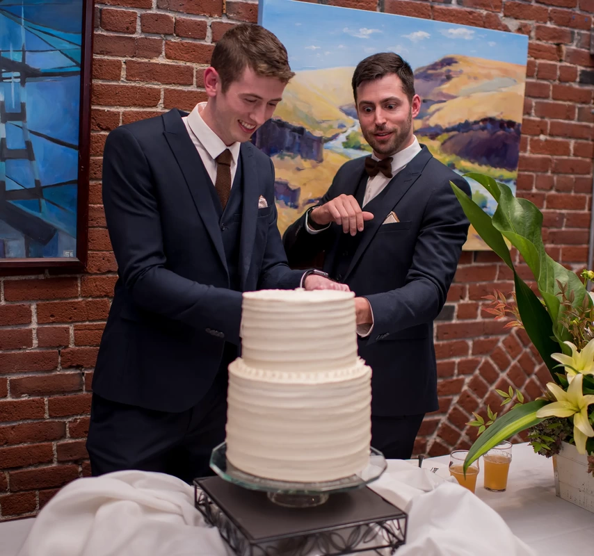 the grooms cut the wedding cake together, one groom smiles while the other groom has a look of shock LGBT Wedding Photographer Robert Knapp at ​Tanner Springs Park in Portland Oregon