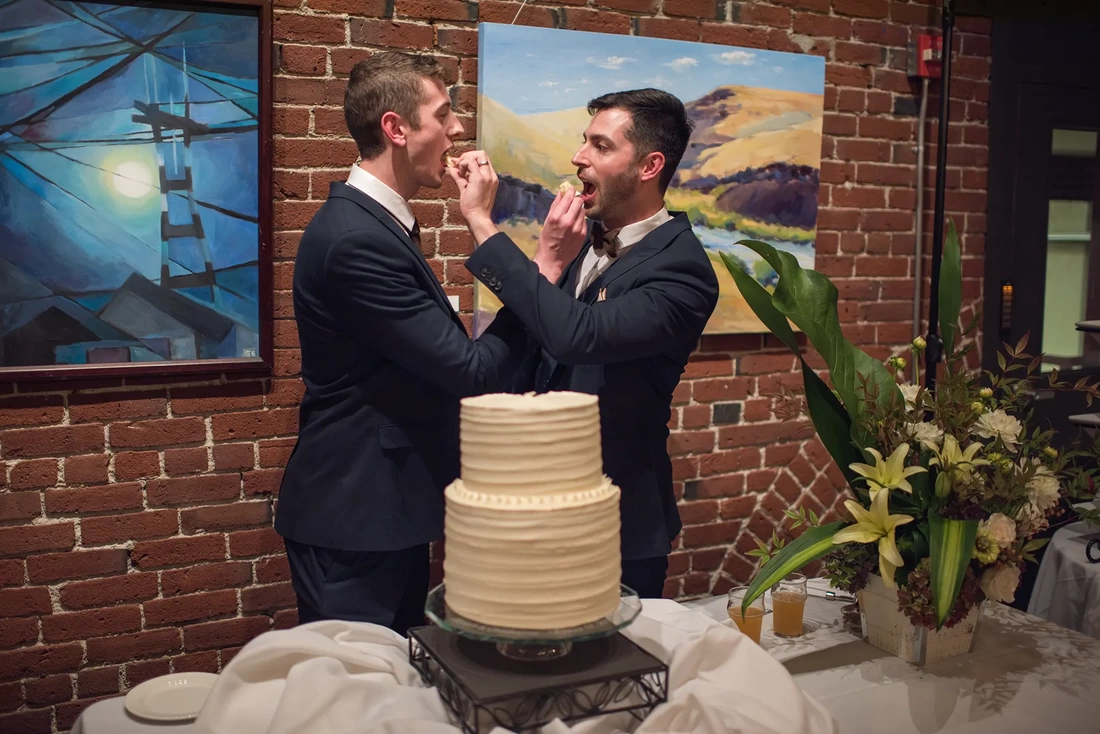 two grooms pass cake to each other after cutting the cake LGBT Wedding Photographer Robert Knapp at ​Tanner Springs Park in Portland Oregon