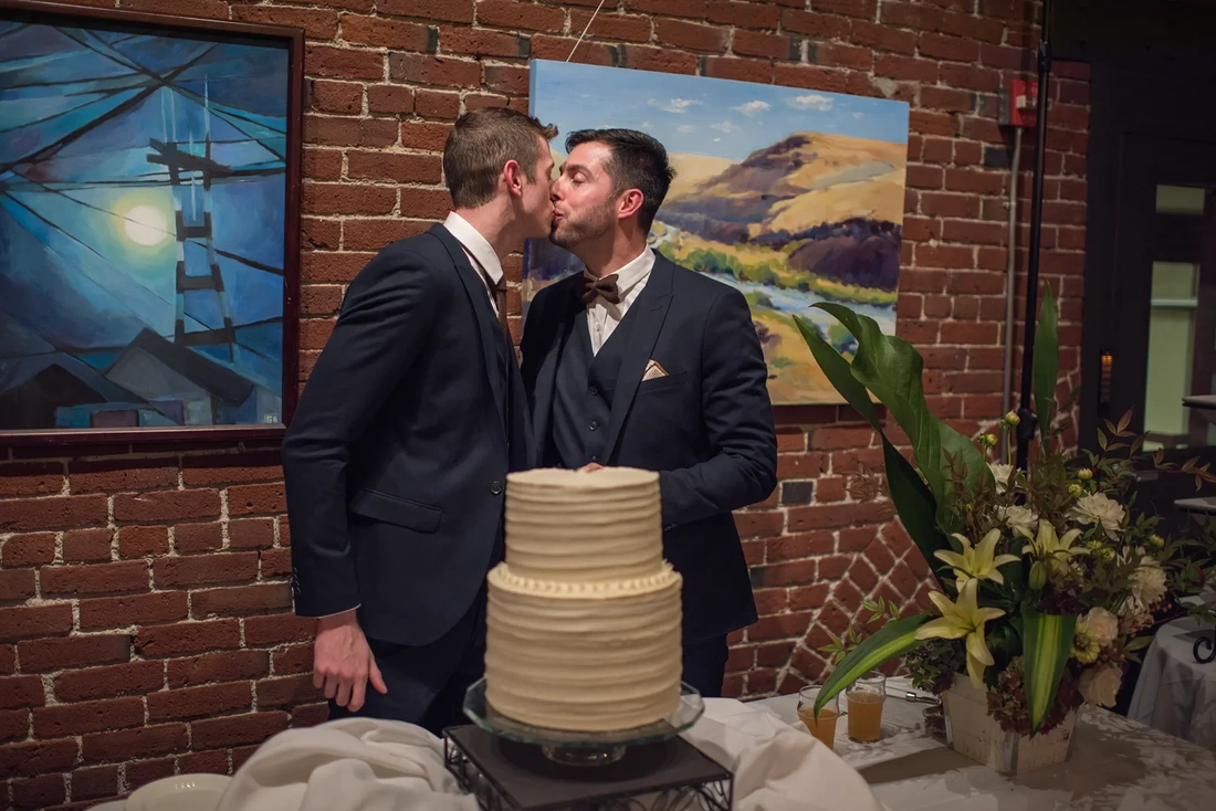 two grooms kiss after cutting the cake LGBT Wedding Photographer Robert Knapp at ​Tanner Springs Park in Portland Oregon