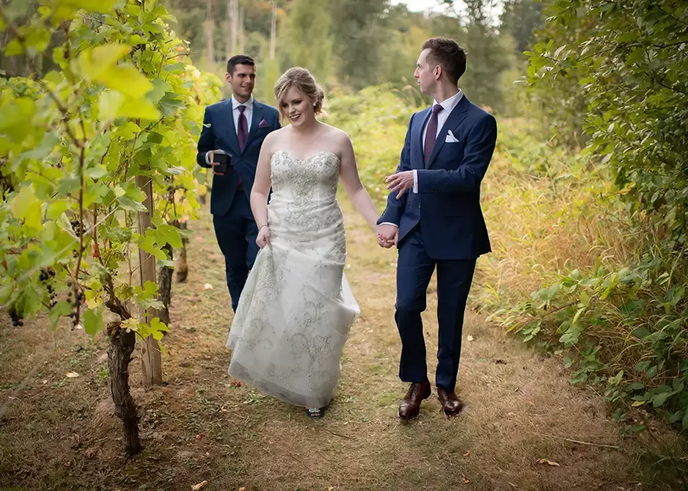 McMenamins Edgefield Weddings with Photographer Robert Knapp the bride and groom walk through a vineyard with a friend. they are all dressed in formal wedding attire of suits and wedding dress.