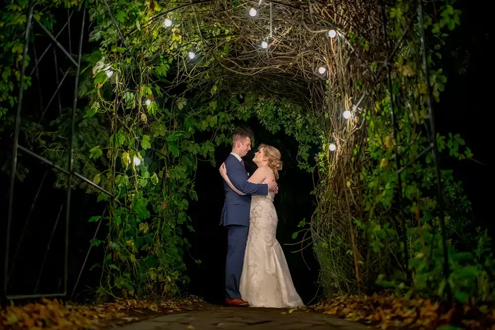 McMenamins Edgefield Weddings with Photographer Robert Knapp bride and groom stand lit by a string of bistro lights under a vine of hops. behind the couple the night is jet black. the bride and groom are lit nicely. the contrast is amazing. 