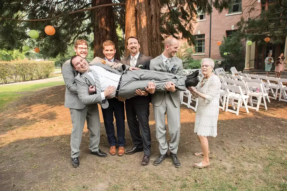 ​McMenamins Grand Lodge Weddings 
from Robert Knapp Photographer a very large groom is lifted by his friends to lay in their arms. A little old woman raises one finger to support the foot. The groomsmen all laugh.