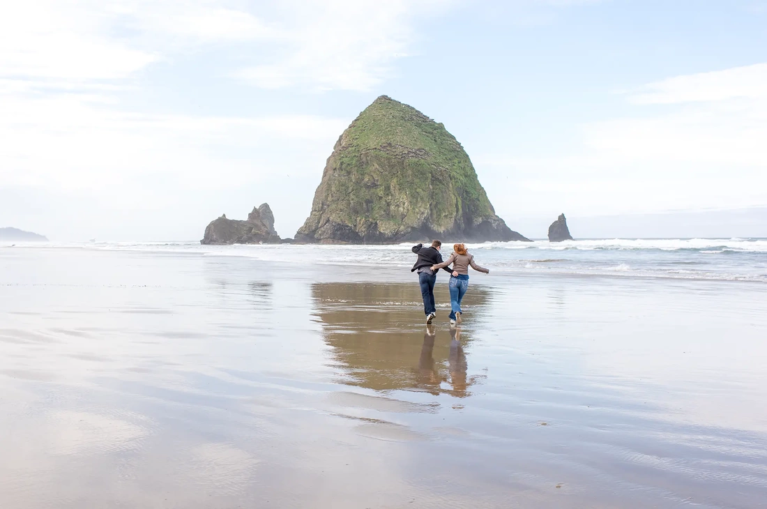 a man and woman run on the beach in front of haystack rock. The beach is wet. The sand reflects them like a mirror. Modern Art Photograph Engagement Photography Portland Oregon​