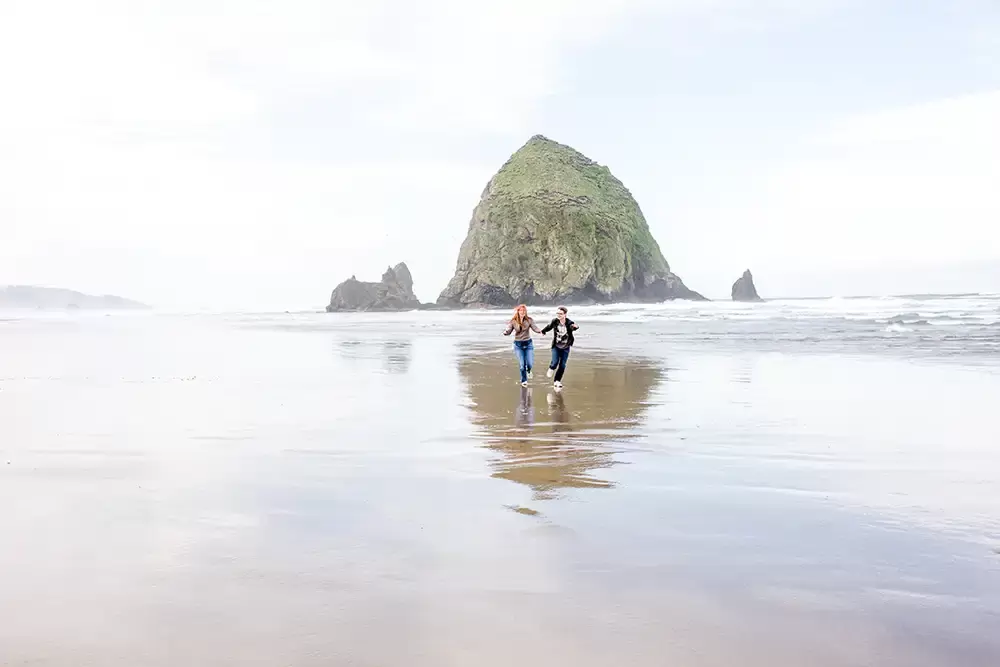 a couple runs to the camera at haystack rock, the wet sand reflects their image. Modern Art Photograph 
Engagement Photography Portland Oregon