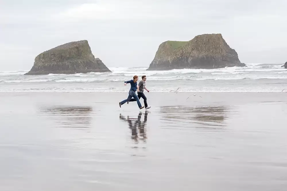 a couple runs along the shore, the waves are close, their step is locked together Modern Art Photograph 
Engagement Photography Portland Oregon