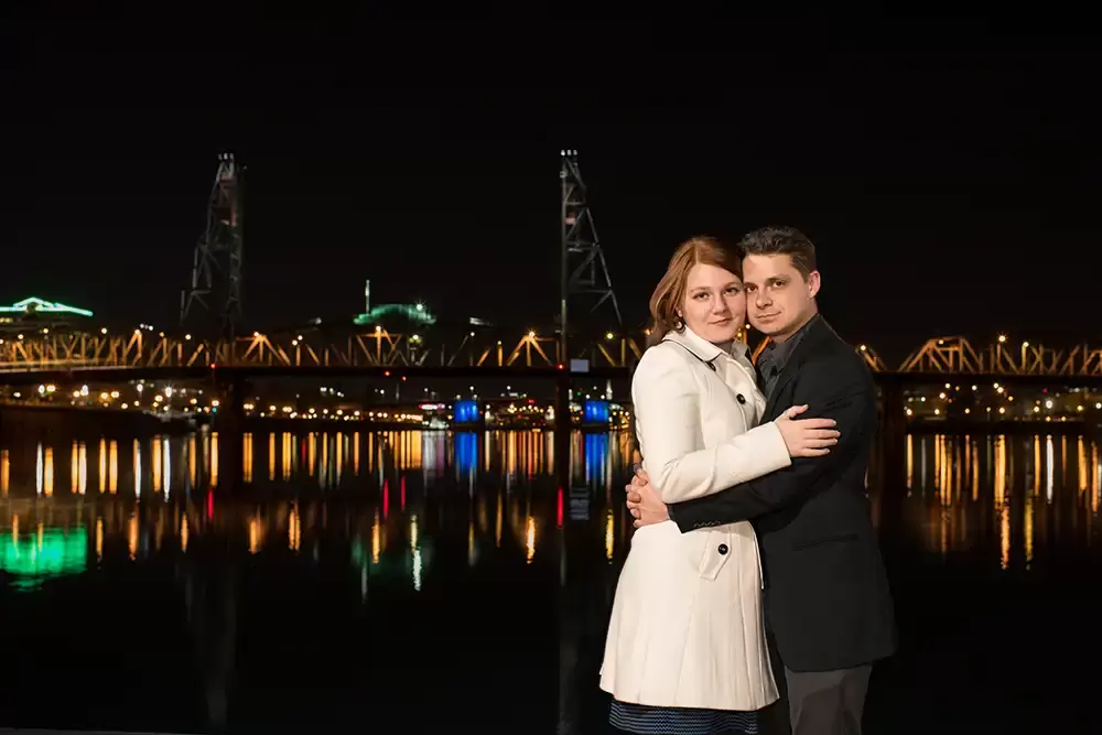 along the skyline a bridge is reflecting across the water against the black of night. A couple holds each other perfectly lit as if it were day, Modern Art Photograph 
Engagement Photography Portland Oregon