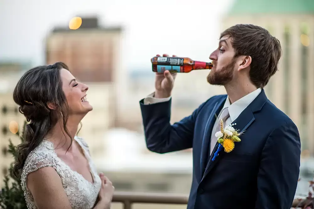 with the city of portland out of focus in the background a bride and groom stand on a balcony. The bride laughs as the groom drinks a beer ​Modern Art Photograph 
Wedding Photographers in Portland
on location at the
Portland Sentinel Hotel