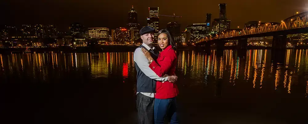 Nighttime Engagement Photos with Modern art Photograph in Portland Oregon, Nighttime engagement photos of a man holding a woman in the dark as the city skyline reflects off the water