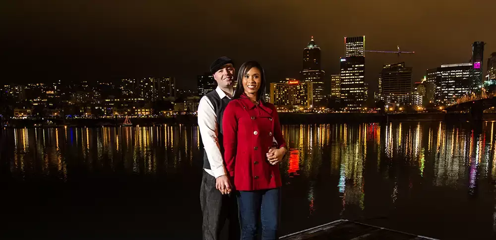 Nighttime Engagement Photos with Modern art Photograph in Portland Oregon, Nighttime engagement photos of a man and woman standing on a dock as the city reflects of the river 