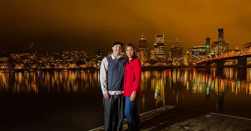 Nighttime Engagement Photos with Modern art Photograph in Portland Oregon. The city reflects golden in the river in these nighttime engagement photos as a couple stands on the dock. 