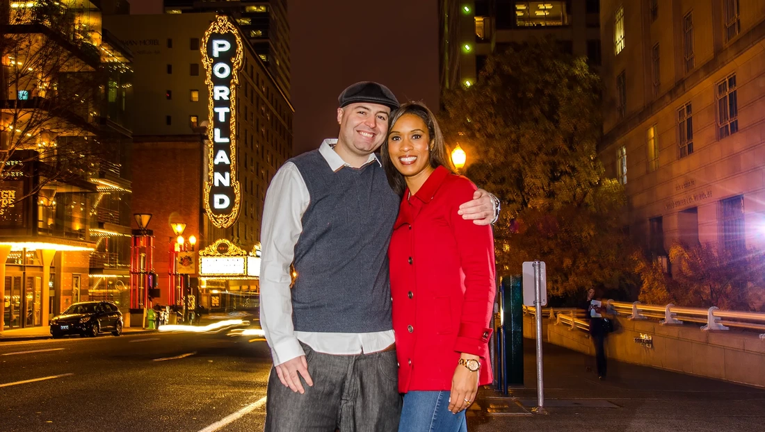 Nighttime Engagement Photos with Modern art Photograph in Portland Oregon, a couple stands on the street with a sparkling lit marquee sign that reads PORTLAND behind them, they have big smiles. 