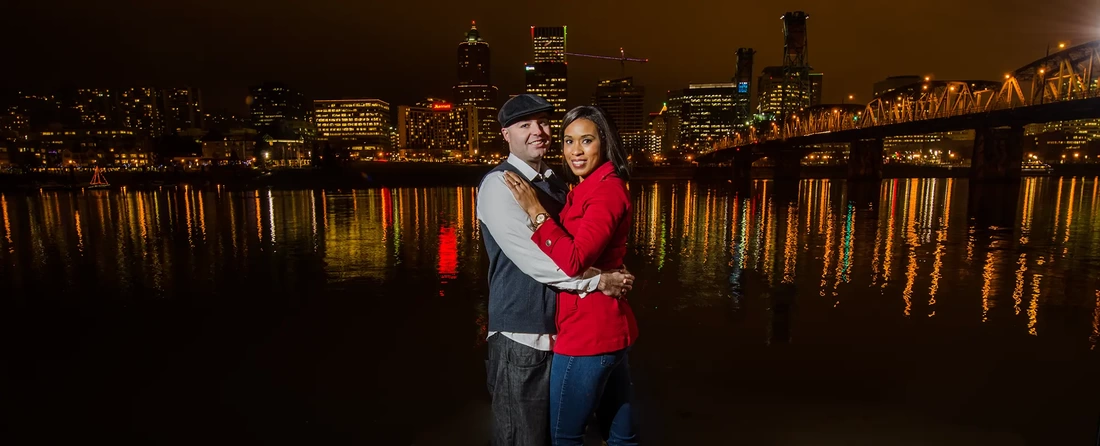 Nighttime Engagement Photos with Modern art Photograph in Portland Oregon a couple holds each other on the banks of the river overlooking the city of portland shining golden in the background 