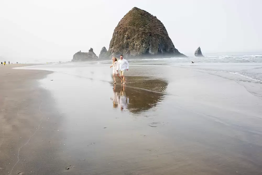 Oregon Coast Engagement Photos 
With 
Modern Art Photograph a couple runs on the beach toward the camera the beach is wet to reflect the horizon of a stone island and the couple 