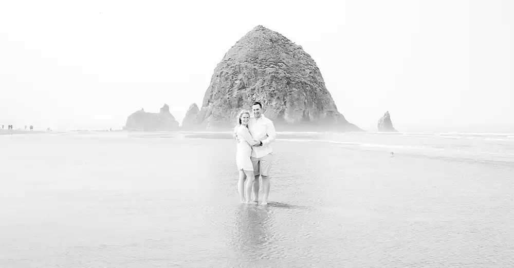 Oregon Coast Engagement Photos 
With 
Modern Art Photograph A couple stands in the morning fog in ankle deep water. Haystack rock is in the distance 