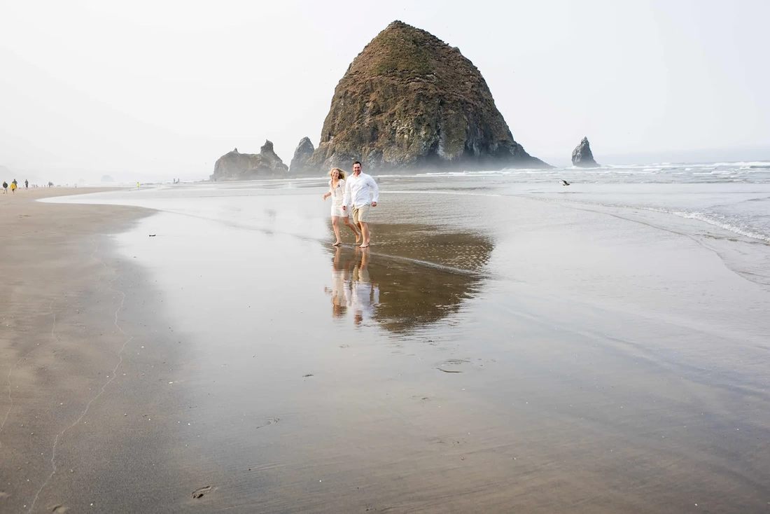 Oregon Coast Engagement Photos With Modern Art Photograph, A couple holds hands and runs through the waves on Cannon Beach